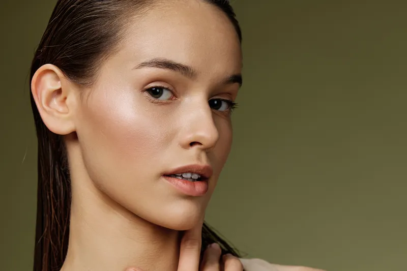 Portrait of a young woman with natural makeup and wet hair, showcasing healthy skin and subtle emotions against a muted green background, perfect for beauty and skincare concepts