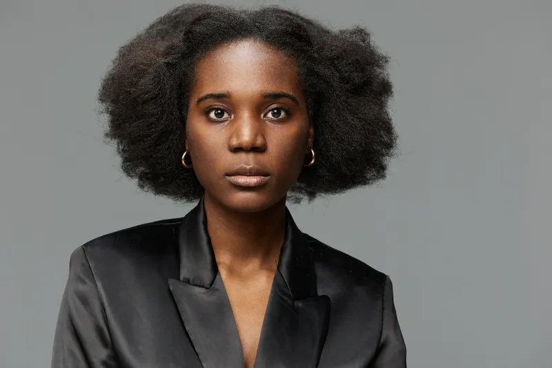 Portrait of a confident Black woman with natural hair, wearing a stylish black satin blazer, showcasing strength and elegance against a minimalist gray background