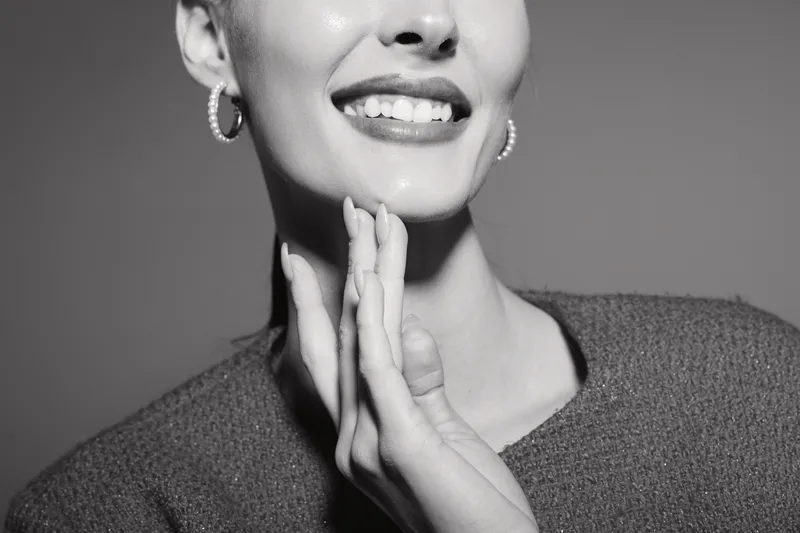 Closeup of a smiling young caucasian woman with light skin touching her chin with hand, wearing hoop earrings and textured sweater. Concept of happiness, beauty, and skincare care routine.