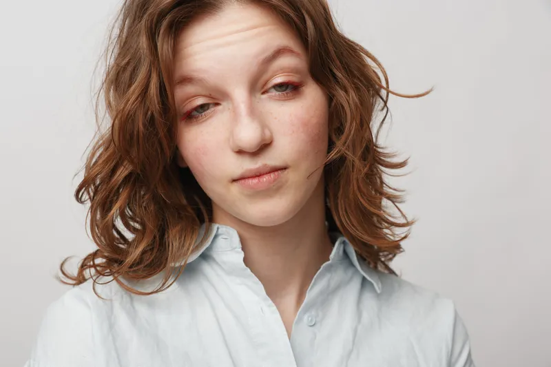 Young woman with curly hair and light blue shirt looks tired and sleepy with half-closed eyes on white background. Concept of fatigue, exhaustion, and low energy in casual portrait.