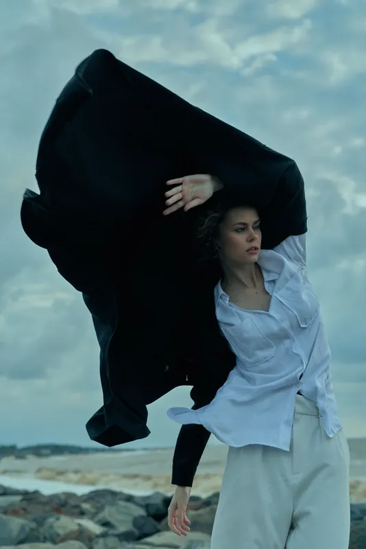 Dramatic fabric cloak sweeps across a windy beach as a woman in a light shirt and loose pants embraces movement against a cloudy sky, creating cinematic fashion and outdoor storytelling
