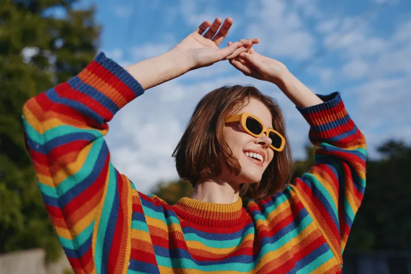 Young woman rainbow sweater yellow sunglasses smiling short bob arms raised outdoor blue sky, joyful summer lifestyle portrait with vibrant striped knit, playful travel vibe and sunlit fashion mood.