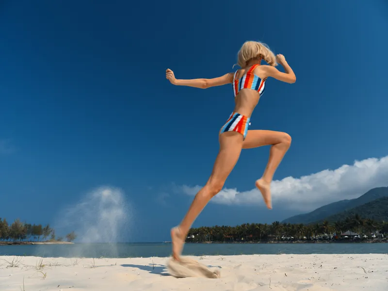 Dynamic action shot of an athletic figure mid jump on a sunny beach, wearing a striped bikini, with powdery sand spray and a vivid blue sky overhead.