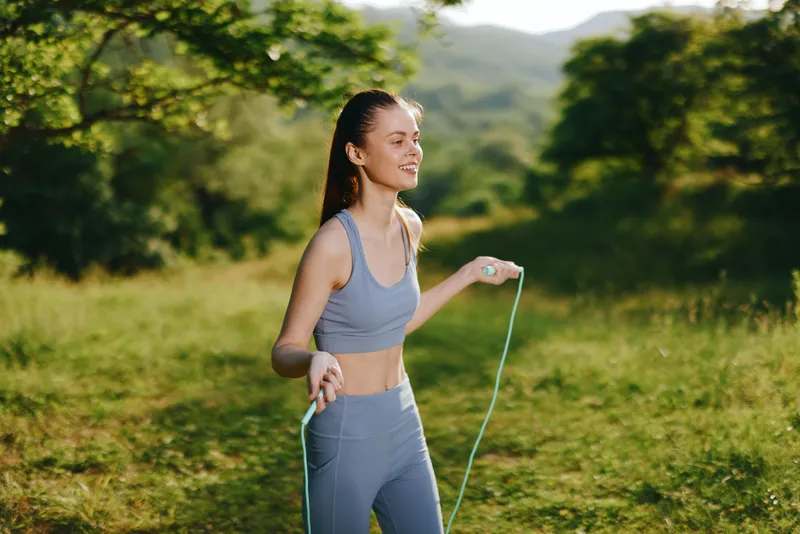 Fit young woman skipping rope in nature, showcasing a healthy lifestyle, wearing sportswear in green and gray colors, surrounded by greenery