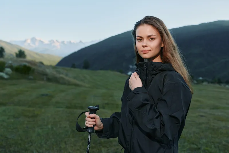 A confident hiker stands outdoors in a scenic mountain landscape. She wears a dark jacket and grips a trekking pole, looking calm, focused, and ready for adventure in nature.