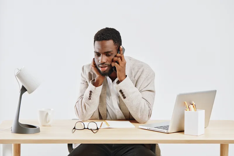 Professional young man with dark skin, beard, and curly hair wearing beige blazer sits at a desk, talking on mobile phone with serious expression, during workday in a modern office setting with minima