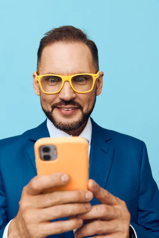 A confident male business professional uses a smartphone, exuding friendly energy against a vibrant blue backdrop. The isolated color field highlights his focused expression and modern style for