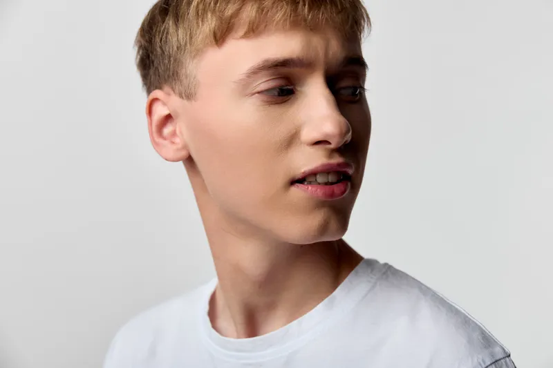 Man in a neutral studio, side profile with a focused expression, wearing a white t-shirt. Soft lighting highlights features against a clean textured background.