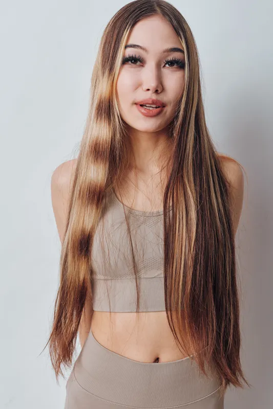 Woman with long straight hair in beige crop top and skirt standing against white background looking at camera with natural expression and minimal makeup.