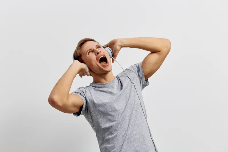 Casual young man with short hair wearing grey t-shirt listens to music on in-ear headphones, expressing joy and excitement with wide open mouth and raised arms, isolated on plain light grey background