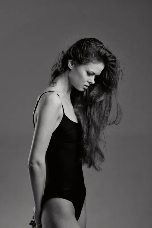 Black and white portrait of a thoughtful woman with long hair wearing a black bodysuit looking down thoughtfully in studio light.