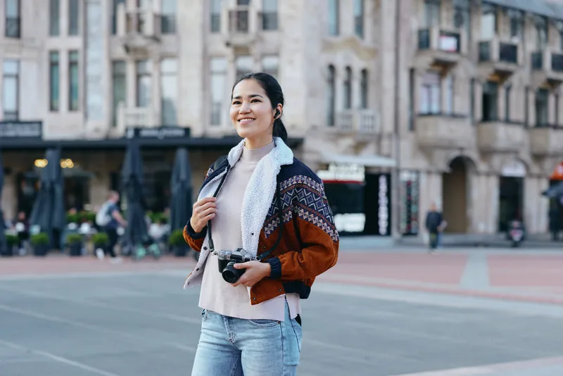 Casual street scene featuring a smiling woman holding a camera in a bustling city square, wearing a patterned knit jacket and jeans, capturing everyday urban life and travel moments.