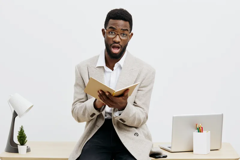 Professional portrait of a surprised man with glasses and dark skin, reading a book at his desk, wearing a beige blazer, minimal office environment, bright natural lighting, focused expression, indoor