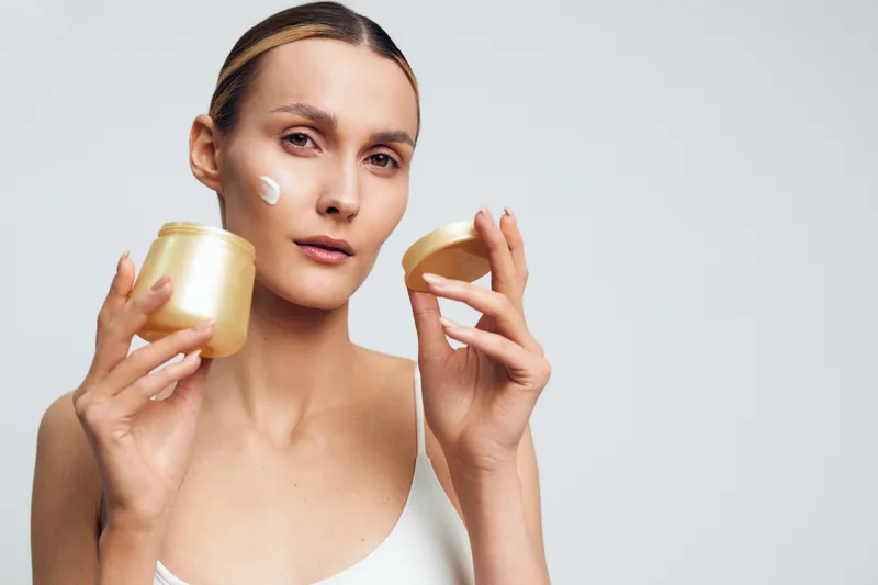 Young woman with natural skin applies face cream from jar on cheek posing isolated on white background, skincare routine, beauty treatment, healthy skin, moisturizing concept.