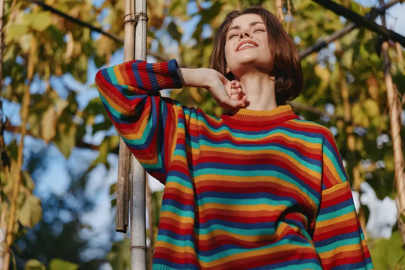 Woman in colorful striped sweater smiling outdoors, short hair and casual outfit, leaning on a bamboo pole under warm sunlight in a garden for joyful lifestyle and fashion portrait