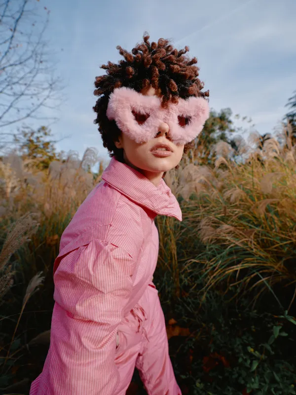 Child wearing a pink outfit and fluffy pink mask in an outdoor meadow, posing with curiosity among tall grasses and a bright sky, conveying imaginative dress up and playful fashion in nature