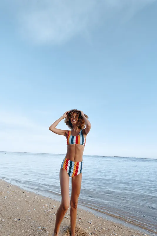 A young woman in a colorful striped bikini stands on a sandy beach with calm sea and clear sky in the background. She holds her hair and smiles enjoying the sunny day.
