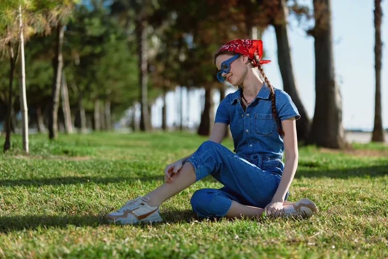young woman sitting on grass in blue denim jumpsuit with red headscarf and sunglasses outdoors on sunny day