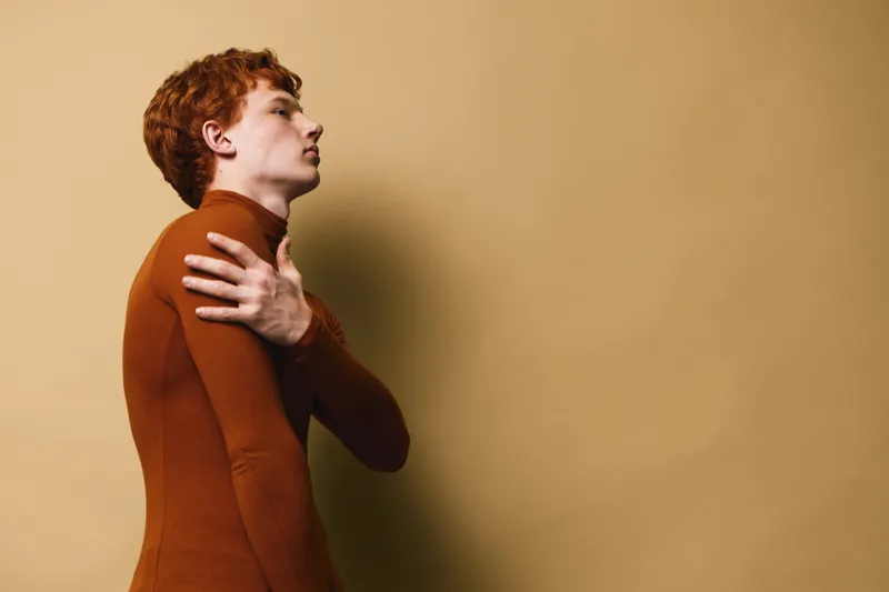Young man with red hair touching shoulder while standing against beige background, profile view showing thoughtful expression and casual warm clothing indoors.