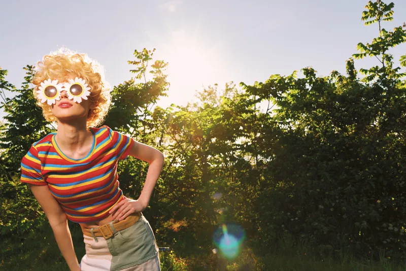 Cheerful woman outdoors, wearing striped shirt and floral glasses, laughing with relaxed posture under bright sunlight, sunlit moment, gentle breeze, lush greenery background, natural colors, retro