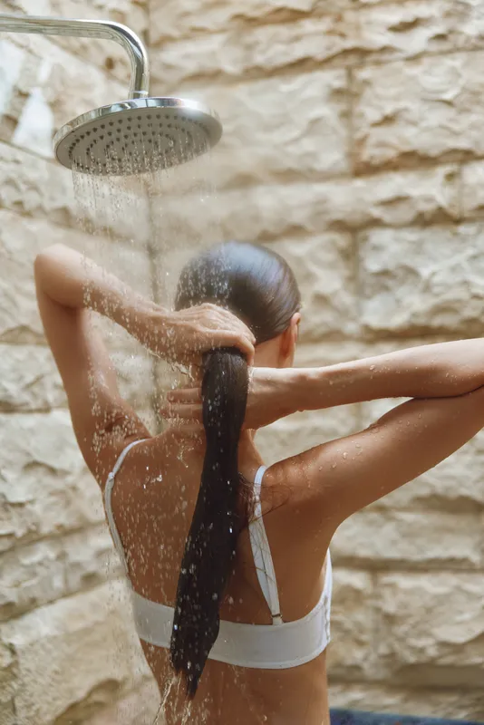 Woman washing hair in outdoor shower with stone wall background, water flowing from showerhead, natural light, wellness and relaxation concept.