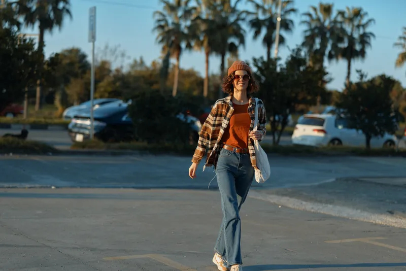 Woman walking casual in denim and beanie, sunglasses on, candid lifestyle portrait in a parking lot with golden hour glow, authenticity and emotional storytelling for mindful living.