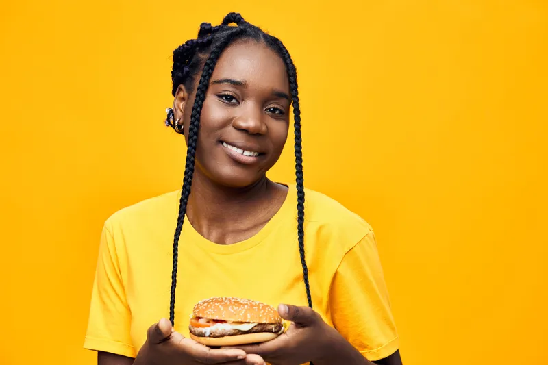 Happy young Black woman holding a delicious burger against a vibrant yellow background, embodying joy and a love for tasty fast food