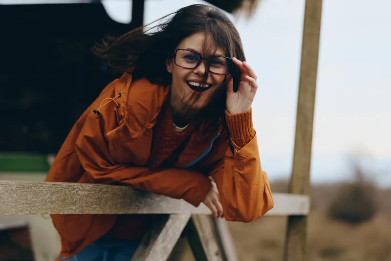 Woman lifestyle outdoor portrait, orange jacket, glasses, laughing with smile, one hand in hair, leaning on wooden fence, blurred background, warm daylight