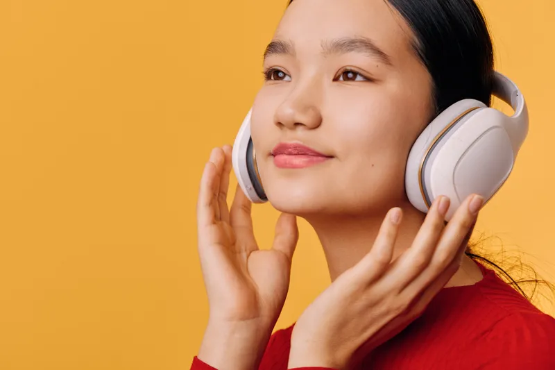 Young woman wearing white headphones smiling and enjoying music on orange background. Female with closed eyes listening contentedly to audio with hands touching ear cups and calm expression.