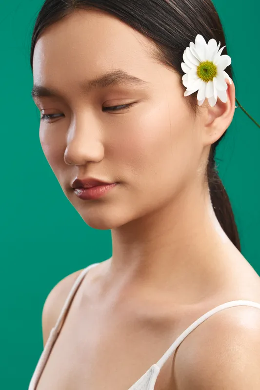 Asian woman with a flower in her hair gazes softly off camera in a studio portrait, presenting calm elegance and natural beauty against a vivid green backdrop.