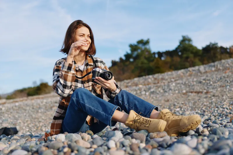 woman, photographer, outdoor, camera, beach, rocks, casual, nature, A relaxed photographer sits on pebbles, capturing a candid seaside moment with a compact camera under a clear sky.