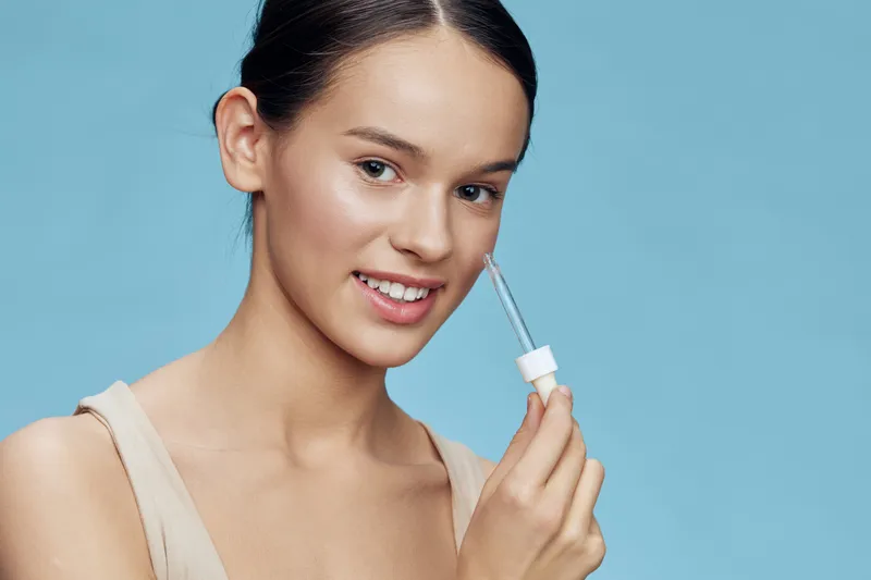 Cheerful young model applying skincare serum on her face against a pastel blue background, promoting healthy skin and beauty routine