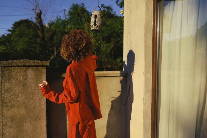 Woman in orange outfit stands on a balcony, back to camera, bathed in sunlight, leaning by the wall for a relaxed urban lifestyle moment outdoors and the soft shadows create calm street mood