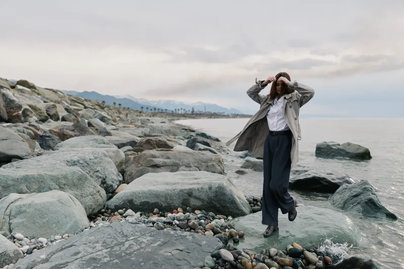 Woman in casual outfit stands on rocky shore by the sea with cloudy sky. Concept for nature, solitude, outdoor lifestyle, and peaceful moments by the water.