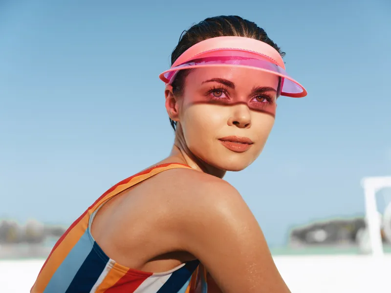 Close-up portrait of a sunlit athlete on a sandy beach, wearing a colorful striped swimsuit and pink visor, glancing over the shoulder with a confident, relaxed expression.