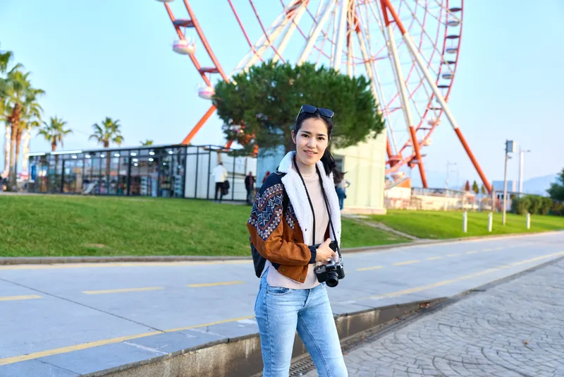 Woman with camera in outdoor amusement park near a ferris wheel, wearing denim jeans and a patterned jacket, capturing candid travel moments under sunny sky.