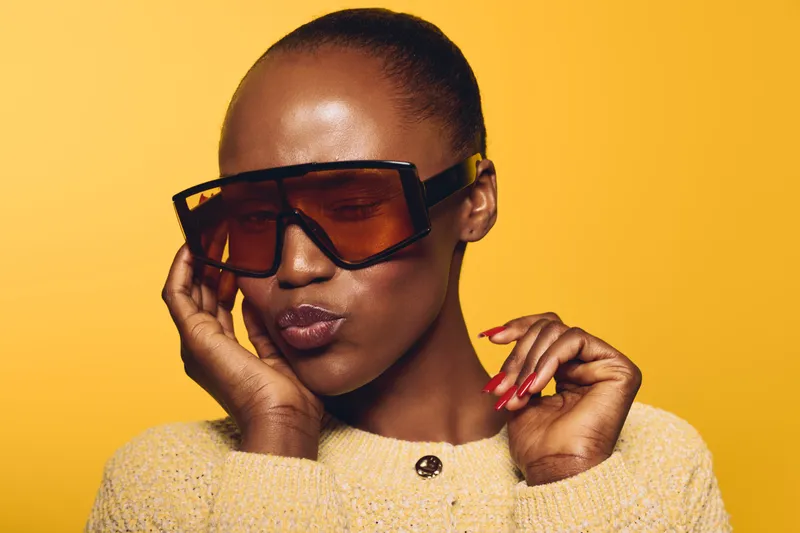 Portrait of a confident black woman with dark skin wearing large brown sunglasses and a beige sweater against a yellow background. Fashion and beauty concept with attitude.