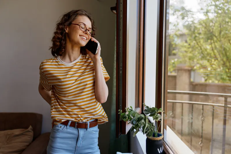 Cheerful woman indoors, talking on phone by a sunlit window, casual striped tee, relaxed pose, plant nearby, soft natural colors, cozy home setting, candid moment