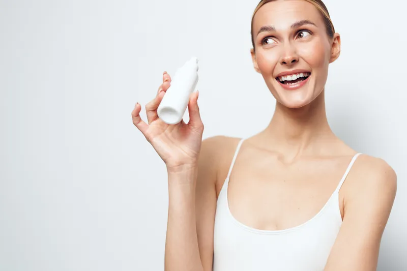 Smiling young woman with blonde hair holds a white cosmetic bottle while looking aside in a bright studio, wearing a white tank top and showing healthy skin and positive emotion.