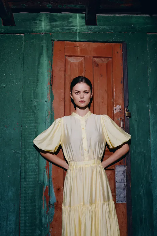 Fashion model stands in a doorway wearing a yellow dress, exuding confidence and poise against a textured green wall, creating a bold editorial portrait for fashion storytelling.