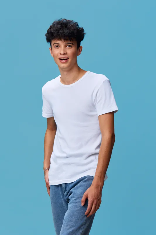 Young man wearing a white t-shirt and blue jeans stands against a blue background with a relaxed and happy expression, showcasing casual style and modern fashion.