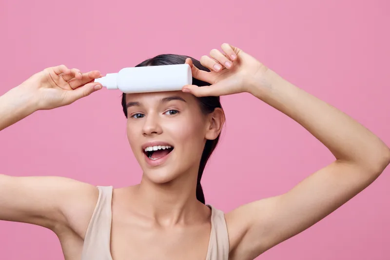 Young woman holding a hair care bottle on her forehead, smiling against a pink background, showcasing a playful expression and promoting hair health