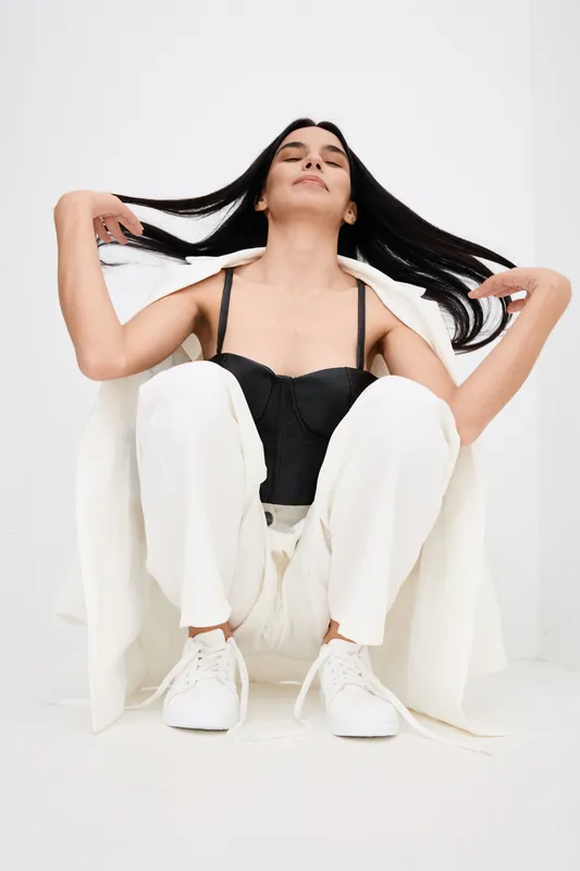 young woman with long dark hair sitting wearing white pants and jacket with black top, playing with hair in bright studio environment