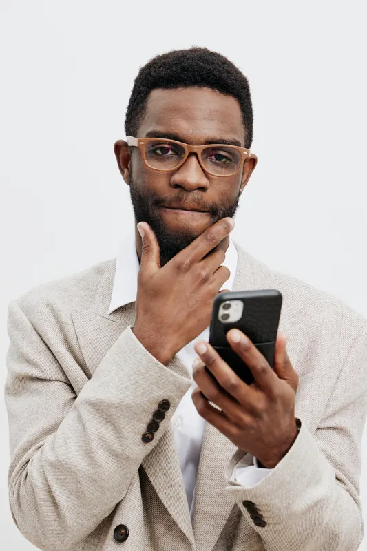 Thoughtful man using smartphone with glasses in hand, dressed in business casual outfit, isolated on a plain light background, studio portrait, modern technology concept, professional communication, s