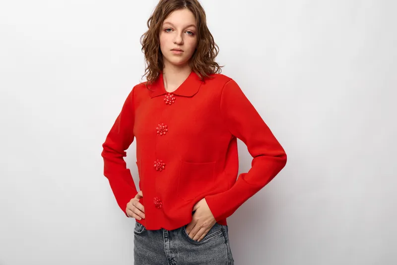 Young woman in red knitted sweater with floral buttons poses with hand on hip against white background. Casual fashion, confident model, autumn style concept for clothing and portrait.