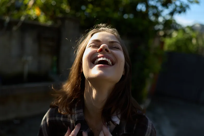 Joyful woman outdoors, head tilted back, laughing with sunlight on her face, capturing a carefree moment in a bright garden setting, casual lifestyle and happiness
