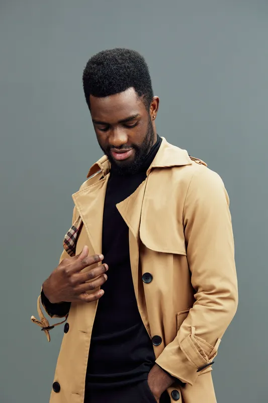 Stylish African man in a beige trench coat with a black sweater, exuding confidence and contemplation against a muted background, reflecting modern fashion trends
