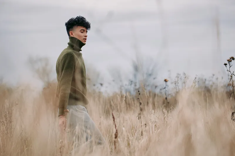 A contemplative young man walks through tall dry grass wearing a textured green sweater, captured with soft natural light to convey simple authenticity and quiet credibility in a candid outdoor