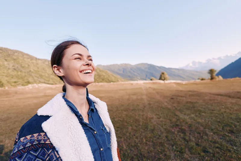 Woman smile portrait in meadow with mountains, outdoors nature scene, happy young adult enjoying fresh air and sunshine, wearing a cozy jacket and blue shirt in rural landscape.