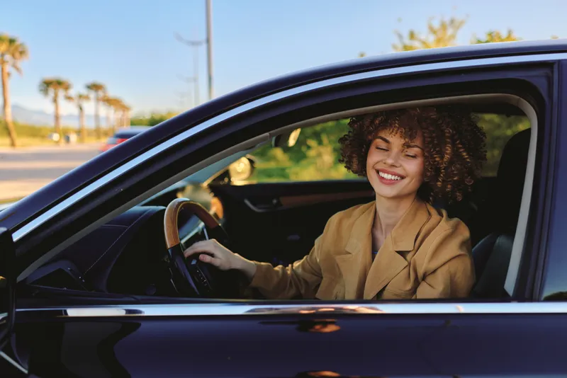 Woman in casual blazer drives a car, smiling, enjoying a sunny daytime drive, lifestyle portrait with open window, urban street scene, warm natural light.